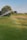 Water sprinklers create a mist over a lush green field, forming a faint rainbow. Trees in the background provide a natural landscape.