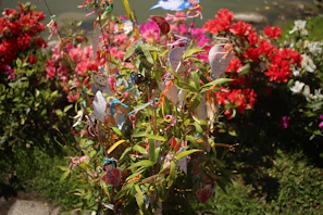 A decorative shrub is adorned with colorful paper ornaments and strings. These ornaments include intricate designs and illustrations, adding an eclectic feel. Surrounding the shrub are vibrant flowers in various shades of red, pink, and white, creating a lush and vivid backdrop.