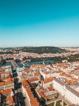 An aerial view of a European city showcasing a dense cluster of historic buildings with red-tiled roofs. A large river runs through the city, crossed by a prominent bridge. In the background, a lush green forested hill contrasts with the urban architecture.