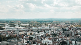 A panoramic view of a cityscape featuring an array of buildings of varying heights and styles, indicating a blend of modern and traditional architecture. In the distance, lush green expanses and forested areas stretch towards the horizon, beneath a sky filled with fluffy, scattered clouds.