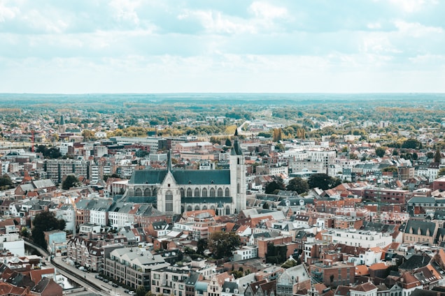 An aerial view of a cityscape with a large, prominent church featuring a tall spire in the center. The surrounding area is filled with densely packed buildings, including houses and apartment complexes. Beyond the city, there are expanses of greenery and a vast, cloudy sky overhead.