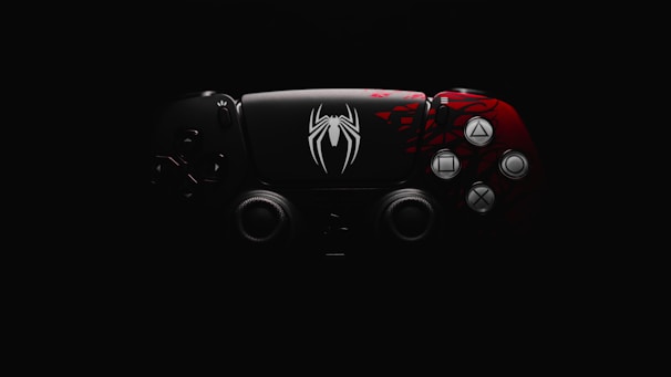 A video game controller features a distinctive design with a black and red color scheme. The left side is predominantly black, while the right side is red with intricate patterns. A prominent white spider emblem adorns the center, adding a thematic element.