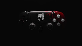 A video game controller features a distinctive design with a black and red color scheme. The left side is predominantly black, while the right side is red with intricate patterns. A prominent white spider emblem adorns the center, adding a thematic element.