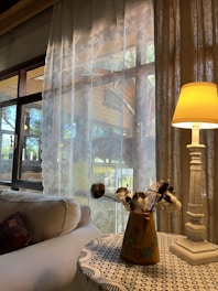 Soft natural light filters through delicate lace curtains, illuminating a cozy corner of a room. A plush sofa with a patterned cushion is positioned beneath the window. Nearby, a small wooden table hosts a vase with cotton branches and a carved white table lamp, creating a charming, homely atmosphere.