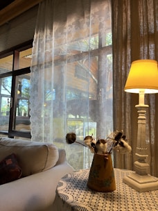 Soft natural light filters through delicate lace curtains, illuminating a cozy corner of a room. A plush sofa with a patterned cushion is positioned beneath the window. Nearby, a small wooden table hosts a vase with cotton branches and a carved white table lamp, creating a charming, homely atmosphere.