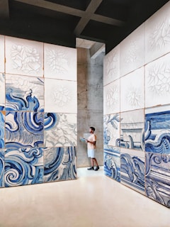 A person stands in an art gallery, surrounded by large panels featuring abstract blue and white swirling designs. The artwork appears to be inspired by water or waves, with intricate patterns. The space is minimalistic, with concrete walls and a clean, open floor.
