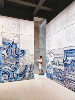 A person stands in an art gallery, surrounded by large panels featuring abstract blue and white swirling designs. The artwork appears to be inspired by water or waves, with intricate patterns. The space is minimalistic, with concrete walls and a clean, open floor.