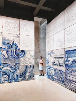 A person stands in an art gallery, surrounded by large panels featuring abstract blue and white swirling designs. The artwork appears to be inspired by water or waves, with intricate patterns. The space is minimalistic, with concrete walls and a clean, open floor.