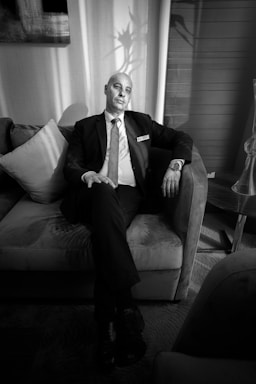 A man in a suit sits on a sofa, appearing relaxed yet formal. The setting is likely a room with soft lighting and a decor that includes pillows and artwork on the wall. Shadows create interesting patterns on the wall, adding a dramatic effect. The man is wearing a name tag and a watch, suggesting a professional environment.