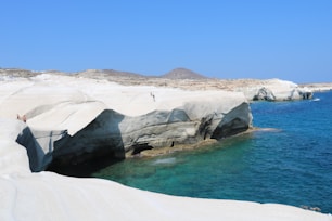 White, smooth cliffs made of volcanic rock sharply contrast with the vibrant blue of the Aegean Sea. A few people are scattered across the rocks, enjoying the sunny weather and the dramatic landscape. The rock formations present unique shapes, carved by natural erosion. In the background, there are barren hills under a clear blue sky.