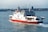 A large ferry with the name 'Red Funnel' painted on the side is navigating through calm waters. The vessel is white with a red base, and there are people visible on the upper deck. The background shows an urban shoreline with buildings and a few cranes, under a clear sky.