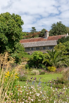 A charming stone house with a red-tiled roof is nestled among lush greenery and colorful wildflowers. Tall trees and various plants surround the building, creating a serene, picturesque setting. The sky is partly cloudy, adding depth to the landscape.