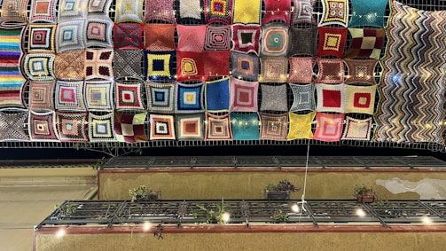 A colorful array of knitted or crocheted squares is suspended above, covering the sky. These vibrant squares feature various patterns and colors, ranging from blues and reds to yellows and more complex multicolored designs. Below the hanging textiles, a balcony with potted plants and a railing is visible. String lights hang slightly below the textile canopy, illuminating the scene against the dark night sky.