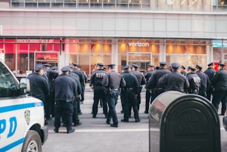 A group of uniformed police officers gathered on a city street in front of a Verizon store. The officers are wearing dark uniforms and caps, and are standing in a semi-circle, engaged in discussion. A police van is parked to the left, and a mailbox is visible in the foreground on the right side.