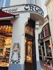 A corner bakery shop with a traditional facade featuring a painted mural of a woman holding bread. The entrance has wooden doors, and inside, there are displays of pastries and baked goods. The exterior is adorned with vintage-style lettering.