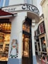 A corner bakery shop with a traditional facade featuring a painted mural of a woman holding bread. The entrance has wooden doors, and inside, there are displays of pastries and baked goods. The exterior is adorned with vintage-style lettering.