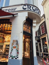 A corner bakery shop with a traditional facade featuring a painted mural of a woman holding bread. The entrance has wooden doors, and inside, there are displays of pastries and baked goods. The exterior is adorned with vintage-style lettering.