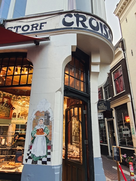 A corner bakery shop with a traditional facade featuring a painted mural of a woman holding bread. The entrance has wooden doors, and inside, there are displays of pastries and baked goods. The exterior is adorned with vintage-style lettering.