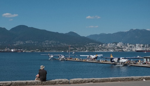 A scenic waterfront view with several seaplanes docked at a pier, surrounded by a vast expanse of water. In the background, a city skyline with numerous buildings is visible, set against a backdrop of forested mountains under a clear blue sky with a few scattered clouds. A person wearing a hat is sitting on a stone wall, facing the water.