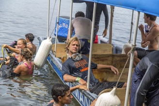 A group of people is interacting in and around a small boat on a body of water. Some individuals are in the water with scuba diving gear preparing for or returning from a dive, while others remain on the boat. One person with goggles rests casually inside the boat.
