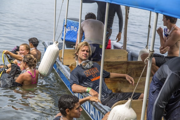 A group of people is interacting in and around a small boat on a body of water. Some individuals are in the water with scuba diving gear preparing for or returning from a dive, while others remain on the boat. One person with goggles rests casually inside the boat.