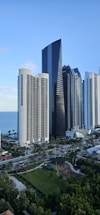A waterfront urban landscape featuring a collection of modern high-rise buildings with unique and varied architectural designs. A large, sleek black skyscraper dominates the skyline, flanked by several other tall buildings. Below, a well-maintained park area with trees and pathways complements the urban setting. The ocean can be seen extending into the horizon under a clear blue sky.