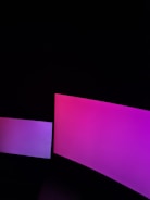 Two screens emit a gradient of pink and purple light in a dark environment. The larger screen on the right displays a more intense pink hue, while the smaller screen on the left has a softer purple tint.