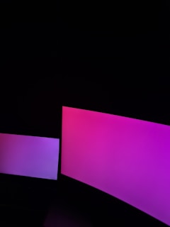 Two screens emit a gradient of pink and purple light in a dark environment. The larger screen on the right displays a more intense pink hue, while the smaller screen on the left has a softer purple tint.