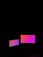 Two digital screens emit a gradient glow of pink and orange hues against a dark background. The larger screen is positioned to the right of the smaller one.