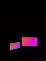 Two digital screens emit a gradient glow of pink and orange hues against a dark background. The larger screen is positioned to the right of the smaller one.