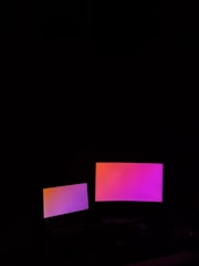 Two digital screens emit a gradient glow of pink and orange hues against a dark background. The larger screen is positioned to the right of the smaller one.
