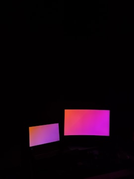Two digital screens emit a gradient glow of pink and orange hues against a dark background. The larger screen is positioned to the right of the smaller one.