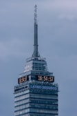 A tall skyscraper with an antenna at the top, featuring a digital clock displaying the same time on two sides. The building has several floors, with a section labeled 'LATINO SEGUROS'. The sky is overcast, giving a greyish backdrop.
