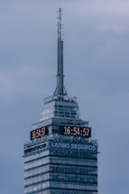 A tall skyscraper with an antenna at the top, featuring a digital clock displaying the same time on two sides. The building has several floors, with a section labeled 'LATINO SEGUROS'. The sky is overcast, giving a greyish backdrop.