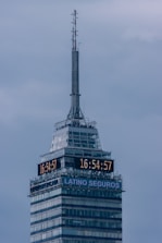 A tall skyscraper with an antenna at the top, featuring a digital clock displaying the same time on two sides. The building has several floors, with a section labeled 'LATINO SEGUROS'. The sky is overcast, giving a greyish backdrop.