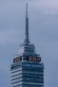 A tall skyscraper with an antenna at the top, featuring a digital clock displaying the same time on two sides. The building has several floors, with a section labeled 'LATINO SEGUROS'. The sky is overcast, giving a greyish backdrop.
