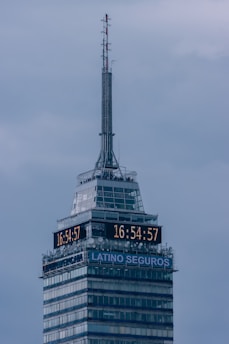A tall skyscraper with an antenna at the top, featuring a digital clock displaying the same time on two sides. The building has several floors, with a section labeled 'LATINO SEGUROS'. The sky is overcast, giving a greyish backdrop.