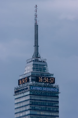 A tall skyscraper with an antenna at the top, featuring a digital clock displaying the same time on two sides. The building has several floors, with a section labeled 'LATINO SEGUROS'. The sky is overcast, giving a greyish backdrop.