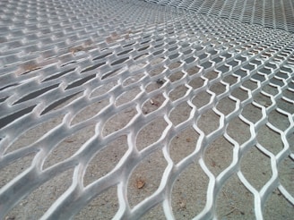 A close-up view of a metallic fence with a hexagonal pattern, extending into the distance. The metallic surface is slightly weathered, and the ground beneath is rough and sandy.