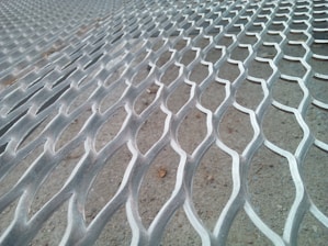 A close-up view of a metal mesh surface with a repetitive, wavy pattern. The background appears to be a concrete floor.