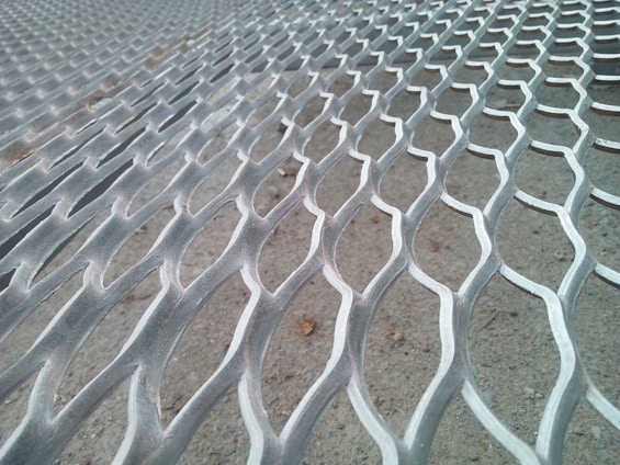 A close-up view of a metal mesh surface with a repetitive, wavy pattern. The background appears to be a concrete floor.