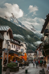 A picturesque street scene in an alpine village, featuring charming buildings with balconies and outdoor seating. The street is bustling with people walking and socializing. In the background, towering snow-capped peaks and evergreen forests create a dramatic and beautiful backdrop, partially shrouded in clouds.
