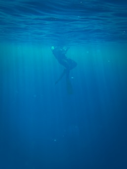A diver is submerged underwater, illuminated by rays of light filtering through the surface. The scene captures the serene and mysterious ambiance of the deep blue sea, with the diver's silhouette partially visible against the water.