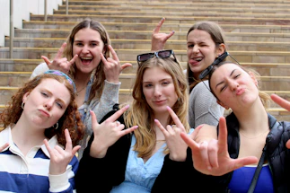 Five young women are standing on outdoor steps, expressing playful and confident gestures with hand signs and facial expressions. They exhibit a sense of camaraderie and enthusiasm, with some wearing sunglasses or casual clothing.