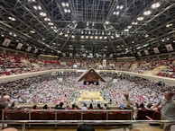 A large indoor arena packed with spectators seated in tiers around a central platform. The platform features traditional Japanese decor, indicating it may be used for sumo wrestling. The atmosphere is bustling, with people filling the seats and many spectators visible in the foreground.