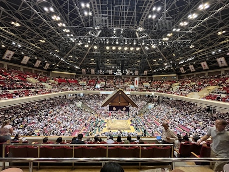 A large indoor arena packed with spectators seated in tiers around a central platform. The platform features traditional Japanese decor, indicating it may be used for sumo wrestling. The atmosphere is bustling, with people filling the seats and many spectators visible in the foreground.
