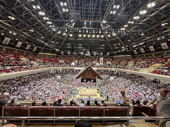 A large indoor arena packed with spectators seated in tiers around a central platform. The platform features traditional Japanese decor, indicating it may be used for sumo wrestling. The atmosphere is bustling, with people filling the seats and many spectators visible in the foreground.