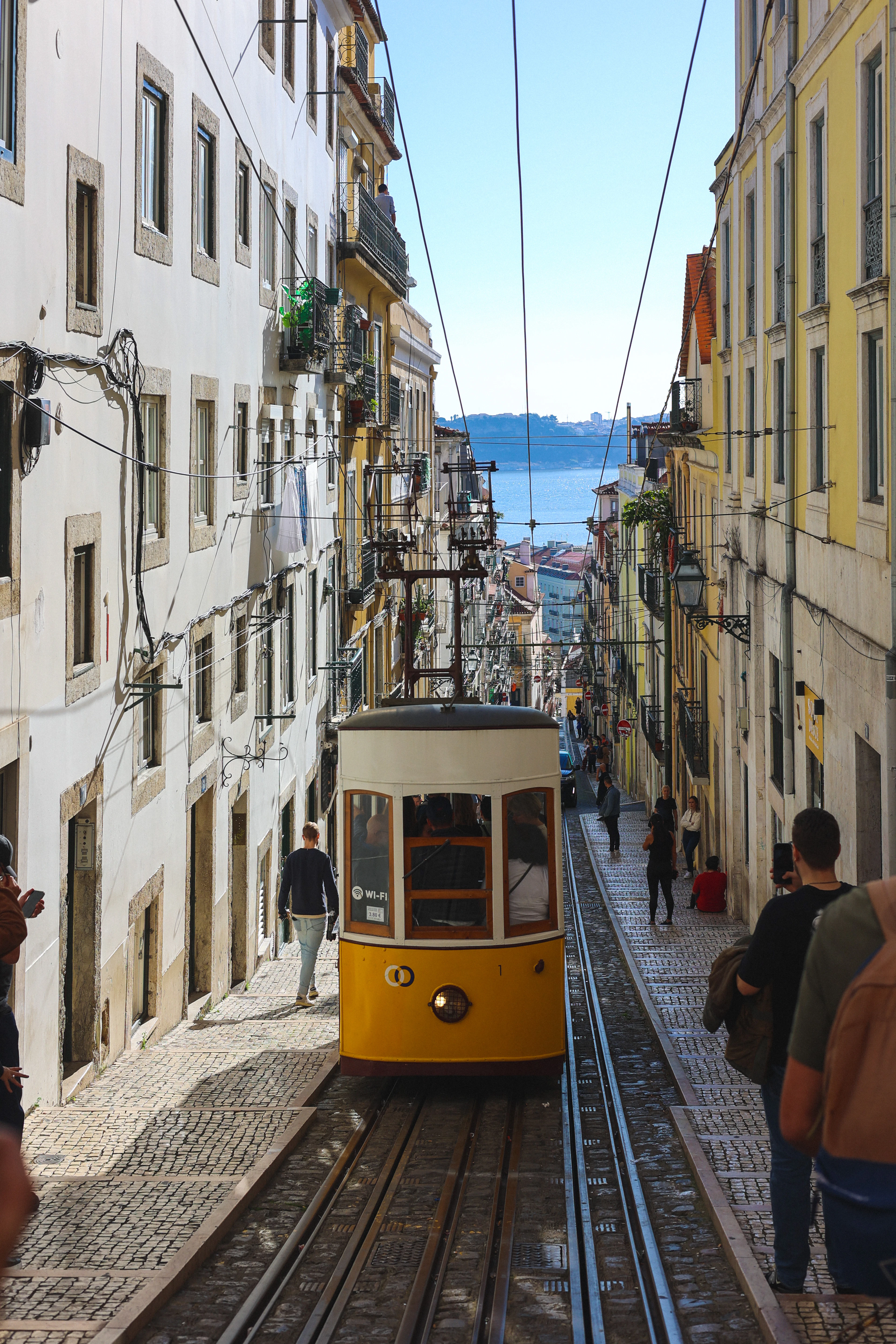 A yellow funicular tram climbs a narrow Lisbon street lined with tiled buildings, the Tagus River beyond.