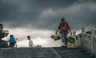 A person is carrying baskets filled with what appears to be durians on a path or bridge. Two children are nearby, and the sky is overcast with dark, threatening clouds. The architectural elements suggest a setting in Asia, with ornate pillars visible.