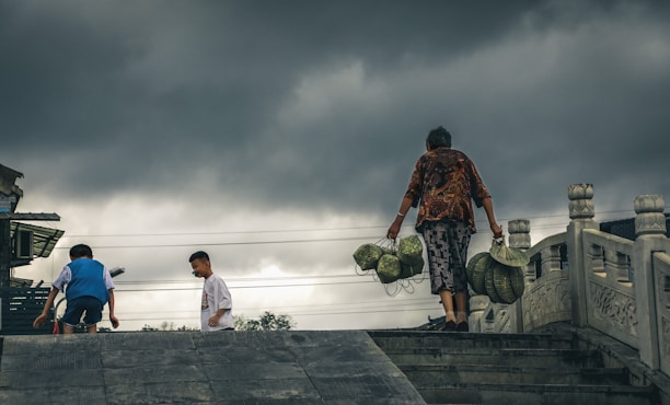 A person is carrying baskets filled with what appears to be durians on a path or bridge. Two children are nearby, and the sky is overcast with dark, threatening clouds. The architectural elements suggest a setting in Asia, with ornate pillars visible.